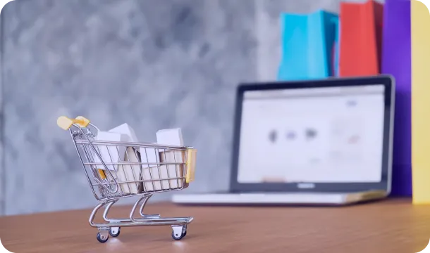 Miniature shopping cart with paper bags on a desk, with a laptop and colorful shopping bags in the background.